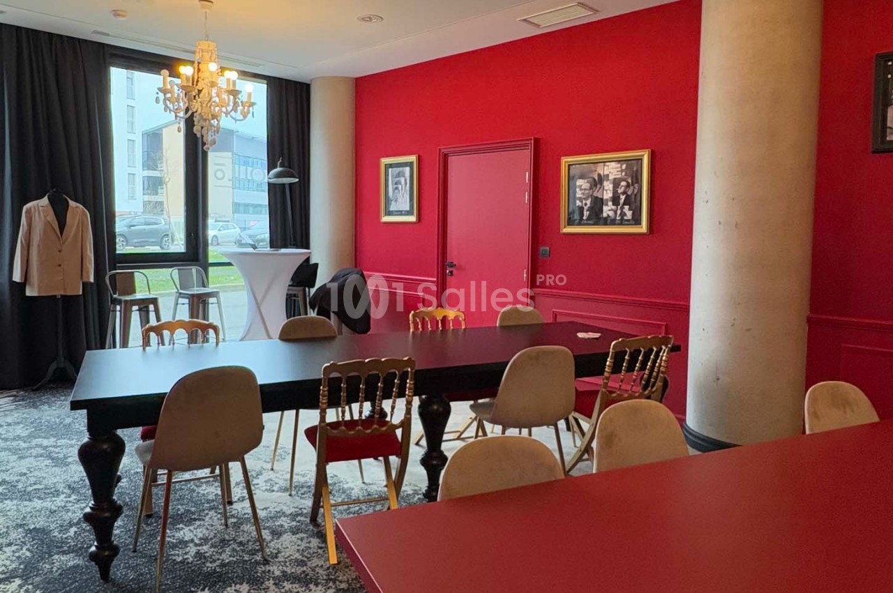 Salle de réunion avec murs rouges, table noire entourée de chaises, lustre et lumière naturelle provenant d'une baie vitrée.