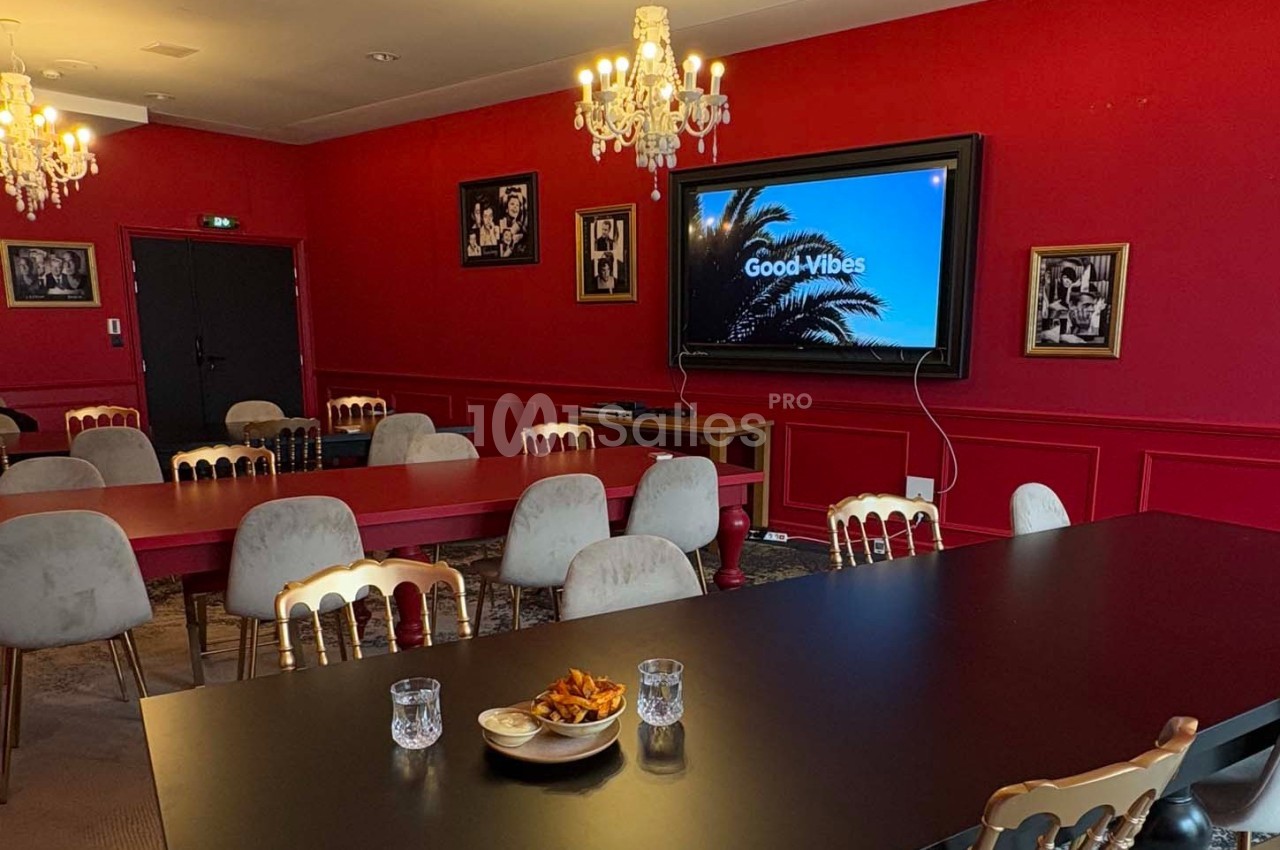 Salle de réunion avec murs rouges, tables noires et rouges, chaises beige, écran affichant ’Good Vibes’, et lustres…