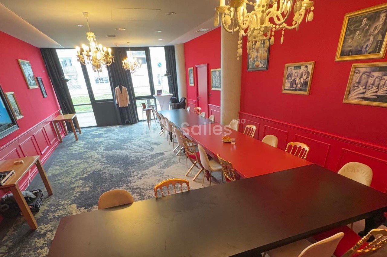 Salle de réunion lumineuse avec murs rouges, tables alignées, chaises variées et lustres suspendus.