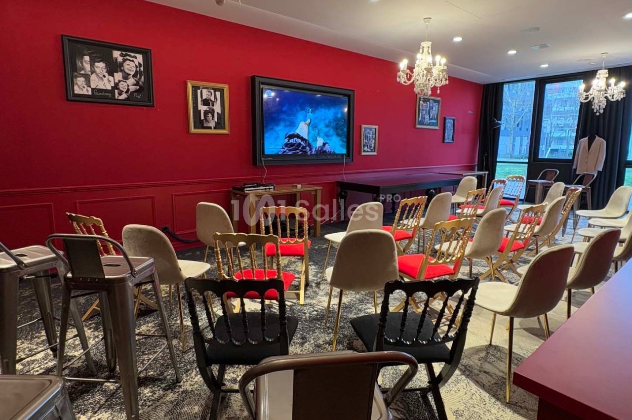 Salle de conférence avec chaises disposées face à un écran, murs rouges décorés de cadres et lustres suspendus.