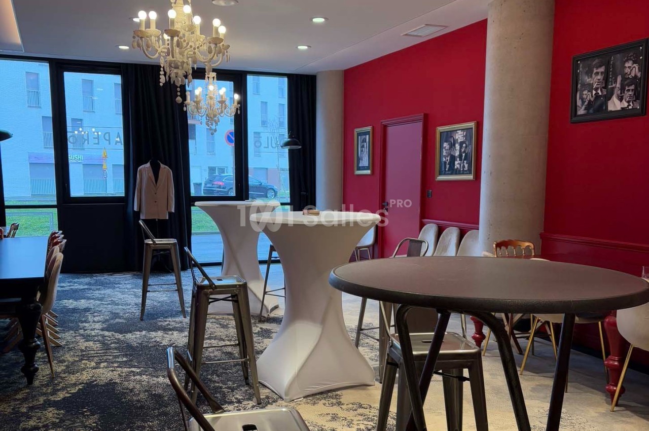 Salle avec murs rouges, tables hautes, chaises métalliques, lustre et photos encadrées, éclairée par des fenêtres.