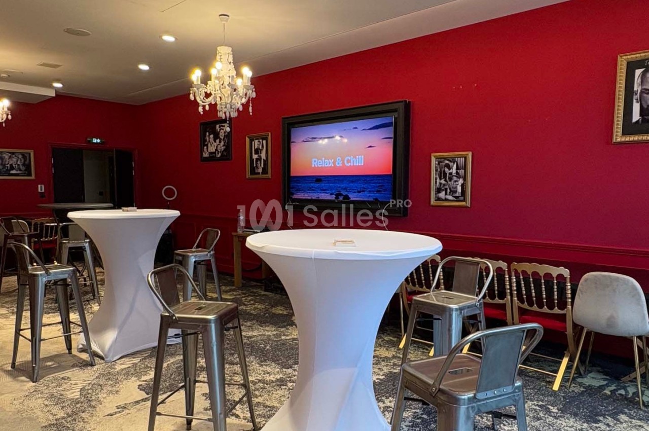 Salle avec murs rouges, tables hautes drapées de blanc, chaises variées et écran affichant ’Relax & Chill’.