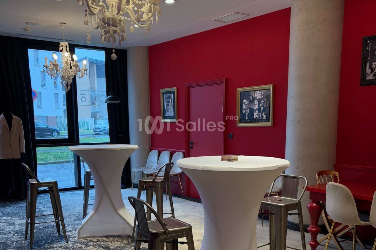 Salle moderne avec murs rouges, tables hautes blanches, chaises métalliques, lustres et cadres décoratifs.