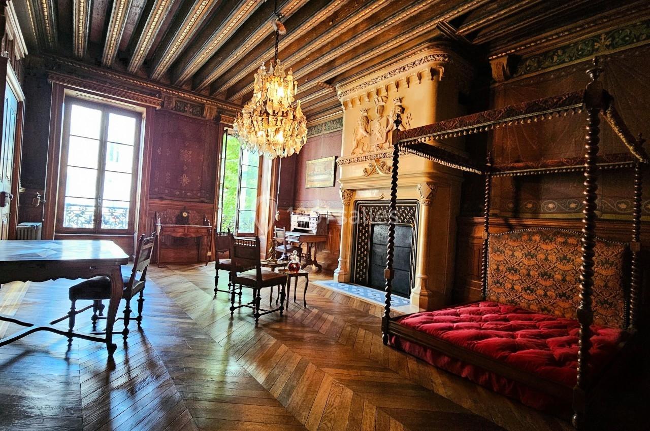 Salle ancienne avec parquet en bois, cheminée sculptée, lustre, lit à baldaquin et mobilier d'époque.