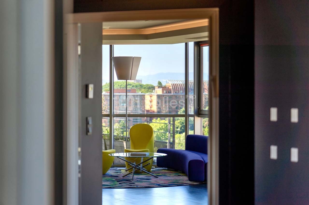 Vue d'un salon moderne avec fauteuils colorés, table basse et grande baie vitrée donnant sur un paysage urbain.