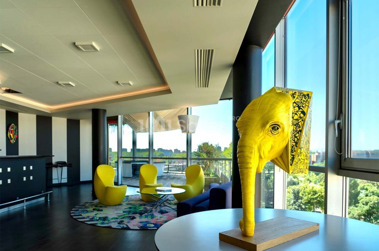 Sculpture jaune en forme de tête d'éléphant sur une table dans un espace moderne avec fauteuils et grandes fenêtres.