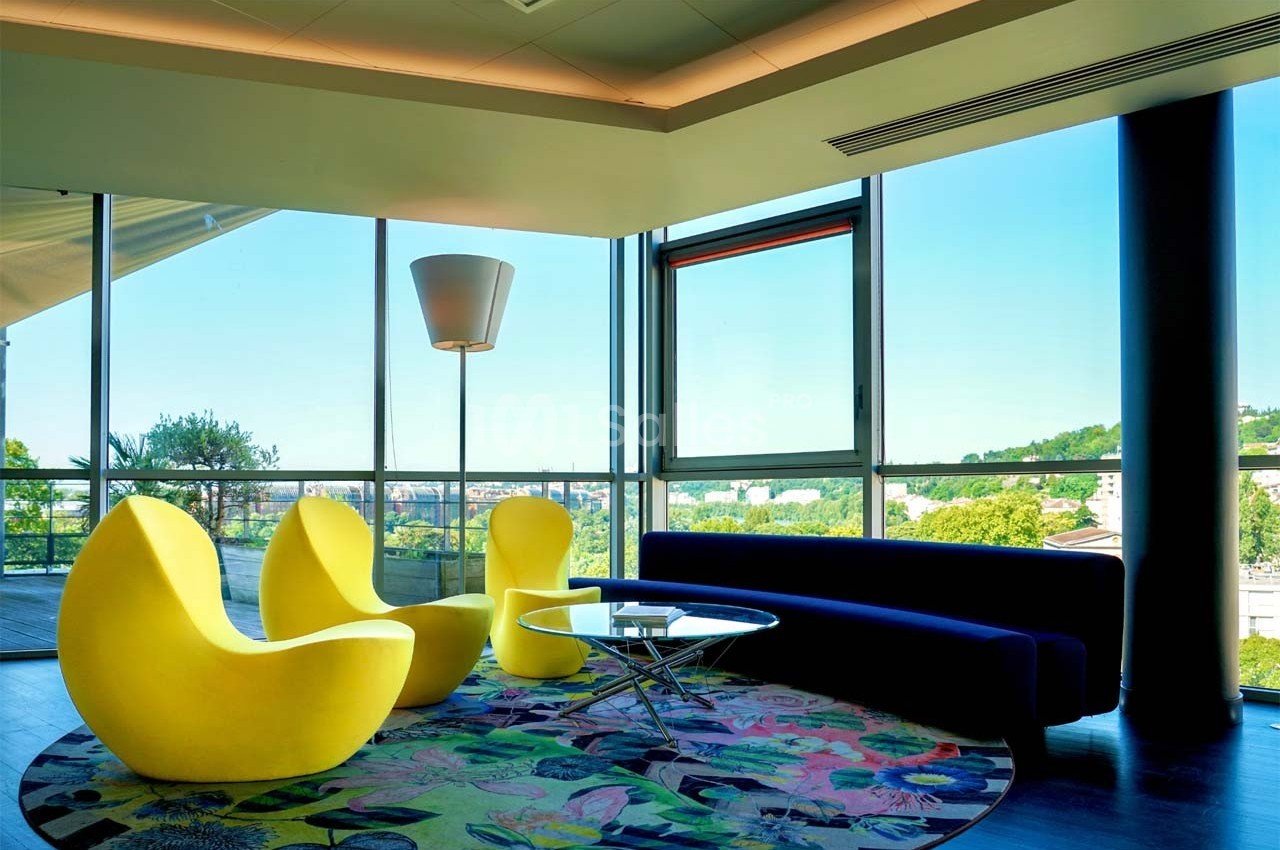 Salon moderne avec fauteuils jaunes, canapé bleu, table basse en verre et grande baie vitrée donnant sur un paysage…