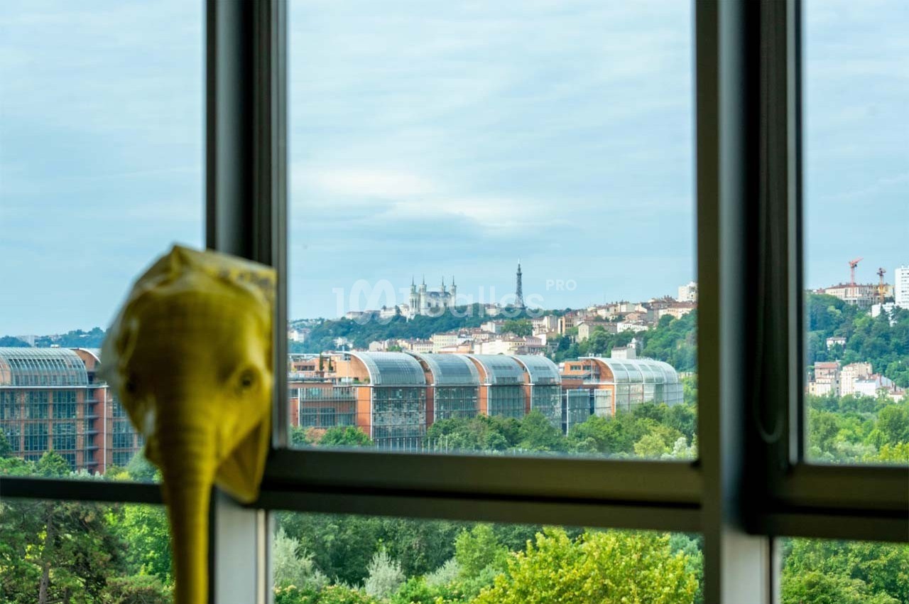 Vue sur des bâtiments modernes, des collines et la basilique de Fourvière à travers une fenêtre, avec une sculpture en…