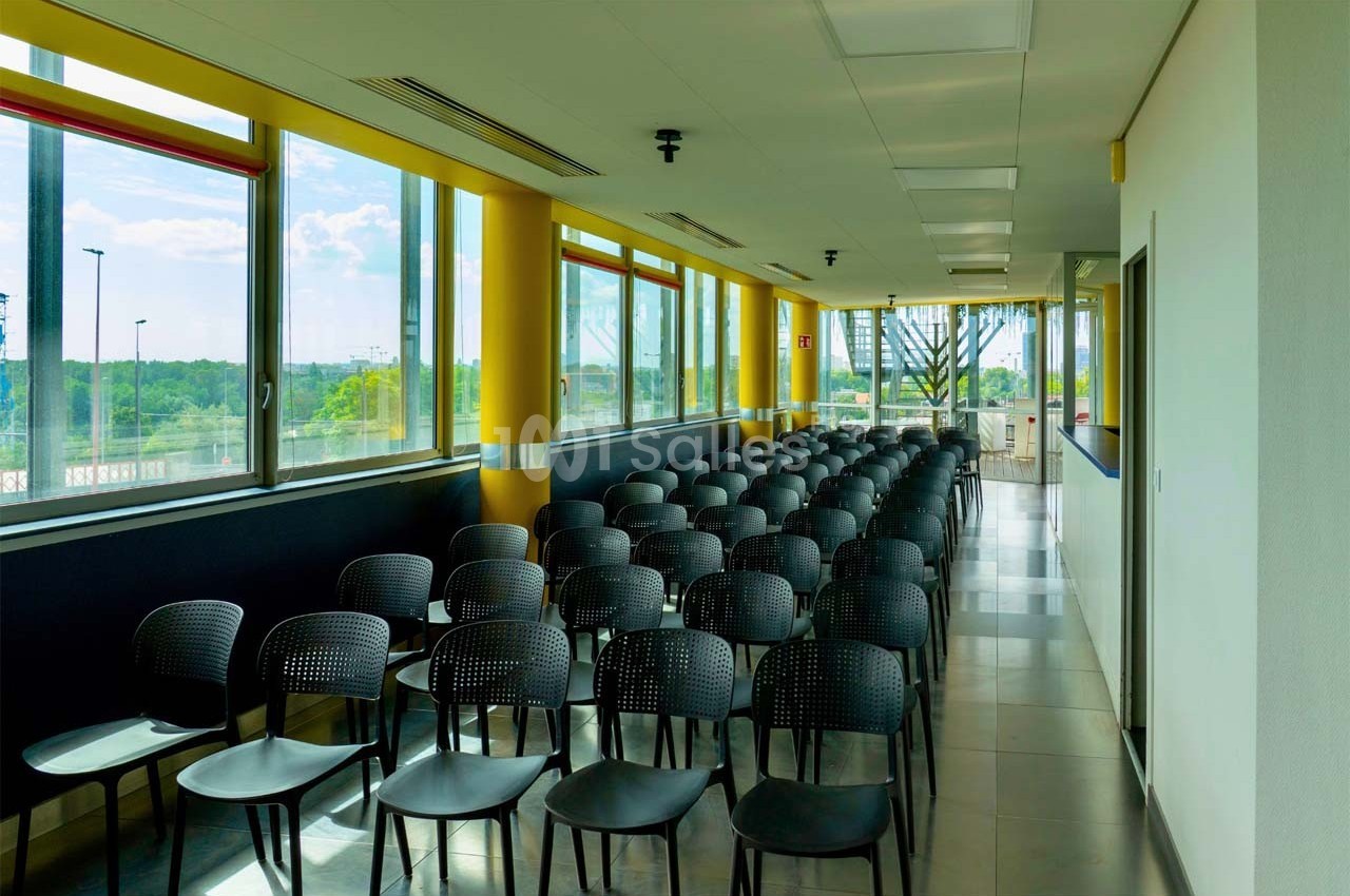 Salle lumineuse avec rangées de chaises noires alignées face à une fenêtre offrant une vue sur un paysage extérieur.