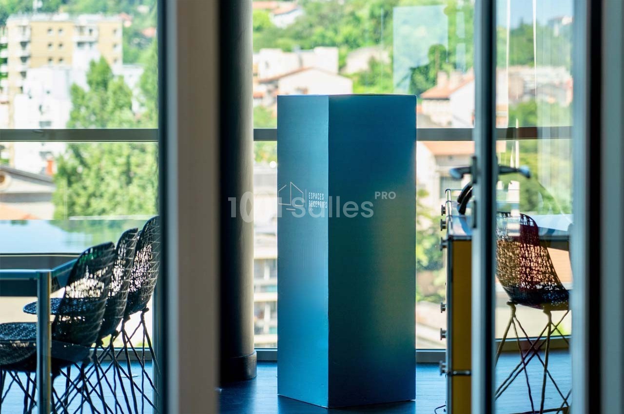 Vue d'une salle de réunion lumineuse avec des chaises en osier et une colonne bleue près d'une baie vitrée.