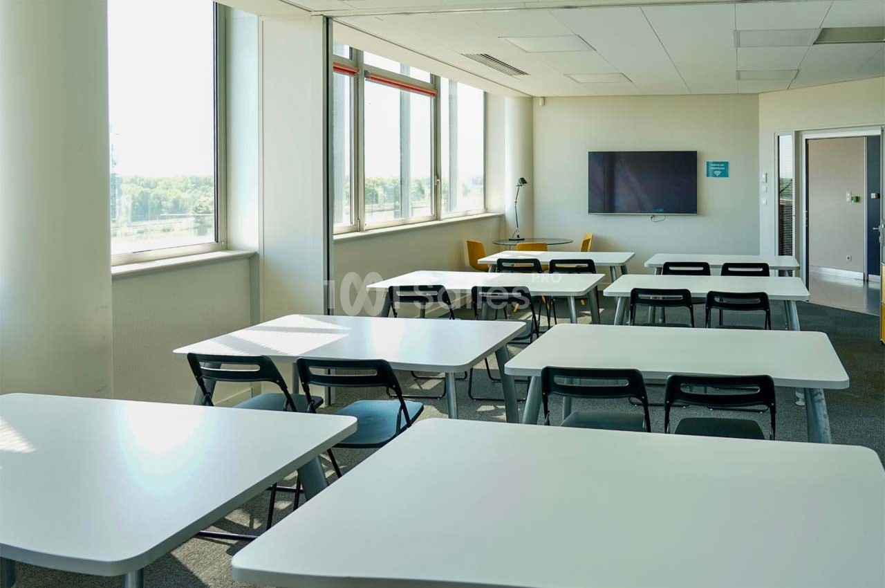 Salle de classe lumineuse avec des tables blanches, des chaises noires, un écran mural et de grandes fenêtres.