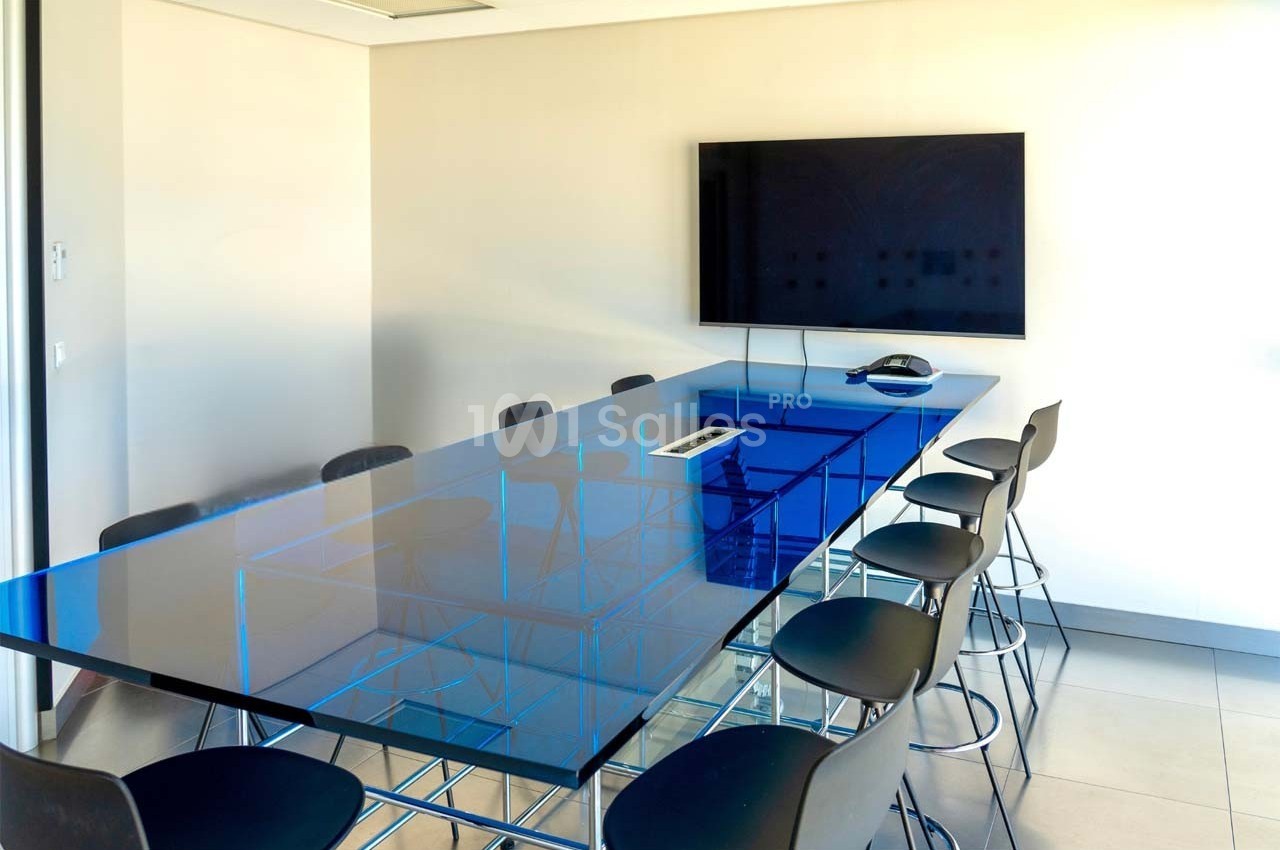Salle de réunion moderne avec une grande table en verre bleu, des chaises noires et un écran mural.