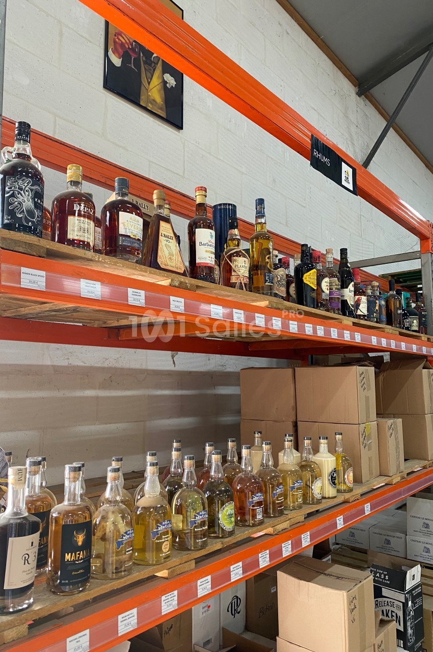 Étagères métalliques contenant diverses bouteilles d'alcool, alignées dans un entrepôt avec des cartons en dessous.