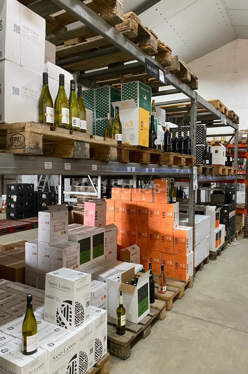 Rayonnages d'entrepôt avec des cartons de vin et quelques bouteilles exposées sur des palettes en bois.