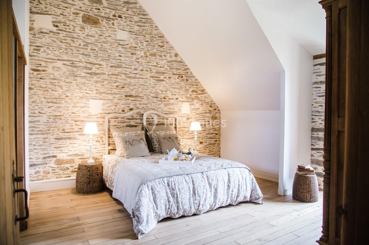 Chambre lumineuse avec un lit double, murs en pierre apparente, parquet clair et deux lampes de chevet allumées.