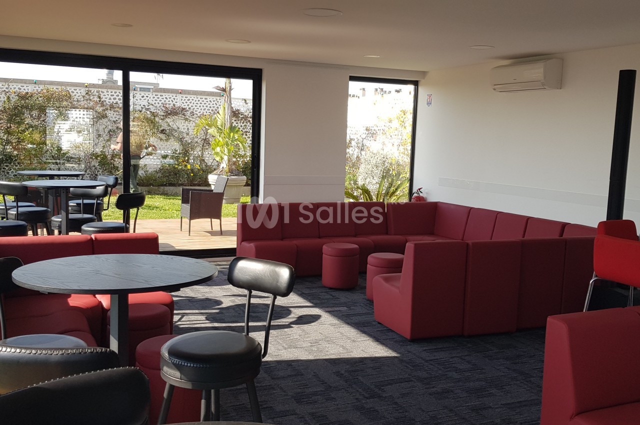 Salle lumineuse avec canapés rouges, tables noires et accès à une terrasse avec des plantes en arrière-plan.