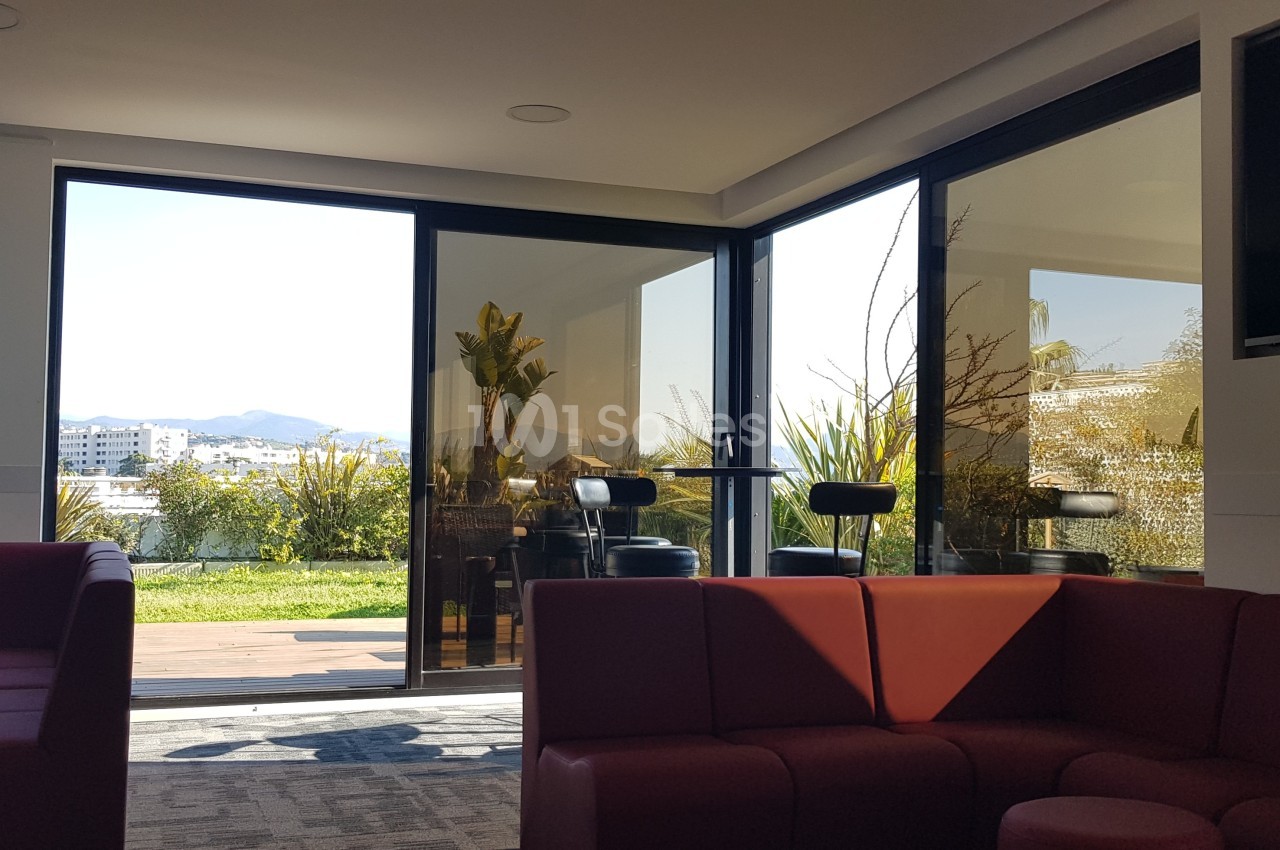 Espace intérieur lumineux avec canapés rouges, grandes baies vitrées donnant sur une terrasse et un jardin.