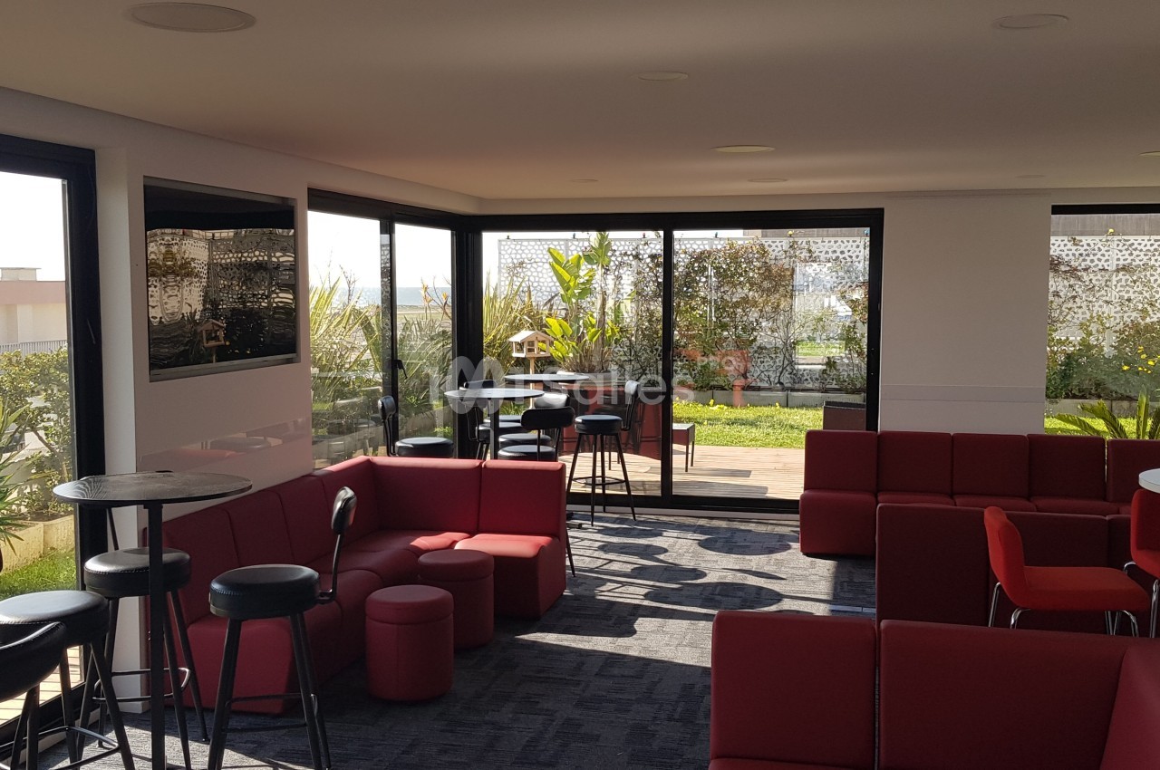 Salle lumineuse avec canapés rouges, tables hautes et vue sur une terrasse végétalisée à travers de grandes baies vitrées.