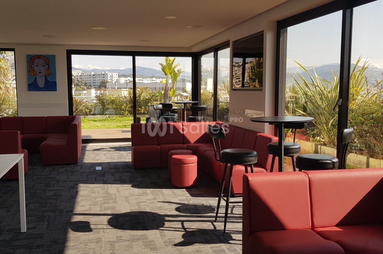 Salle lumineuse avec canapés rouges, tables hautes et grandes baies vitrées donnant sur une terrasse et un jardin.