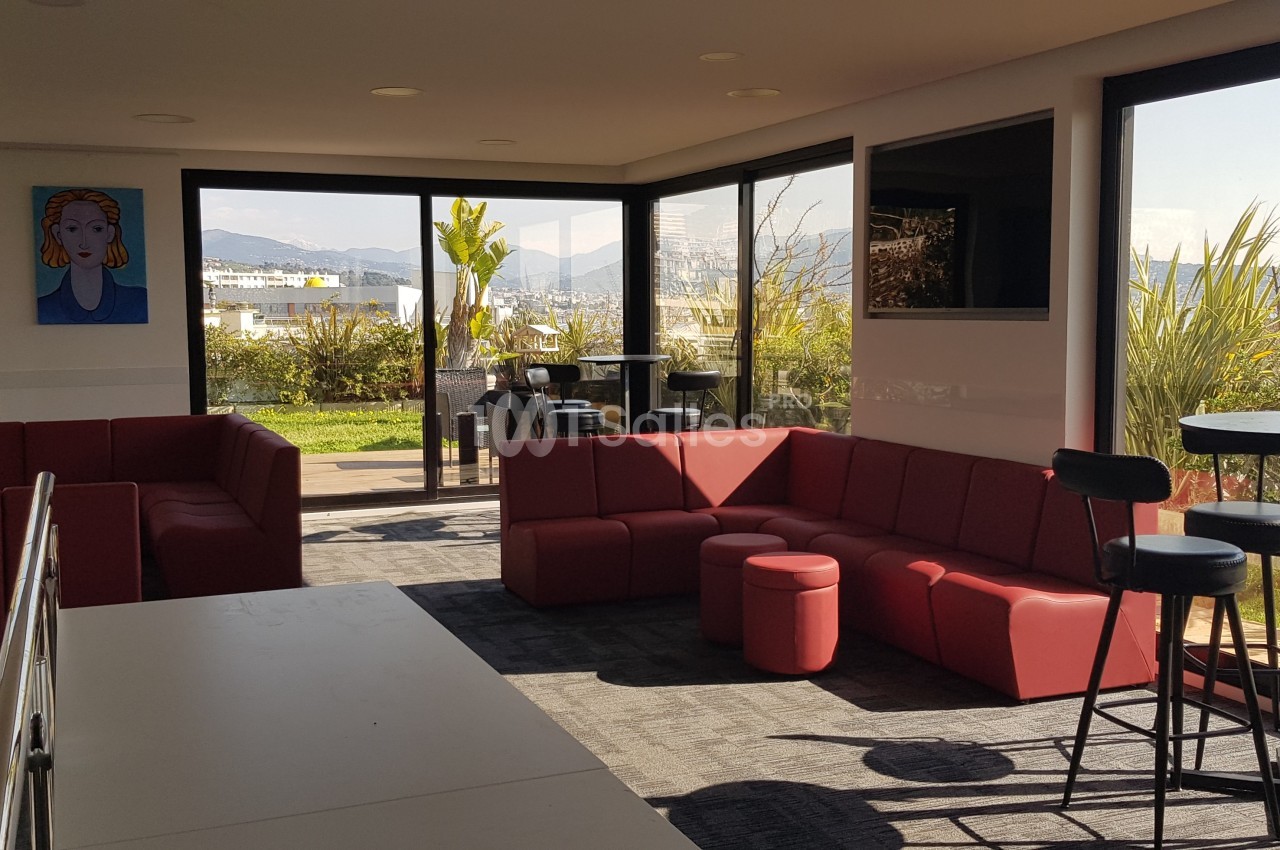 Salle lumineuse avec canapés rouges, tables hautes et vue sur une terrasse arborée à travers de grandes baies vitrées.