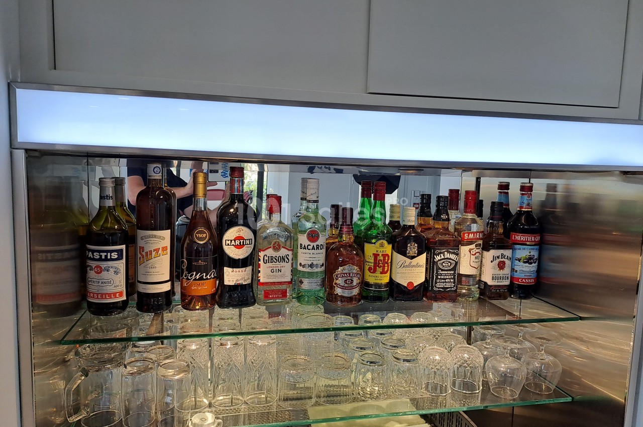 Bouteilles d'alcools variés alignées sur des étagères en verre avec des verres en dessous, dans un meuble éclairé.
