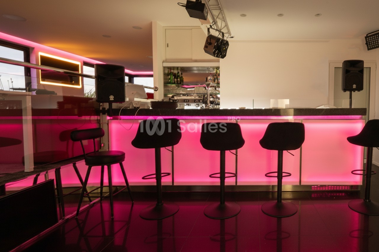 Bar moderne avec comptoir éclairé en rose, tabourets noirs alignés et équipement audio visible en arrière-plan.