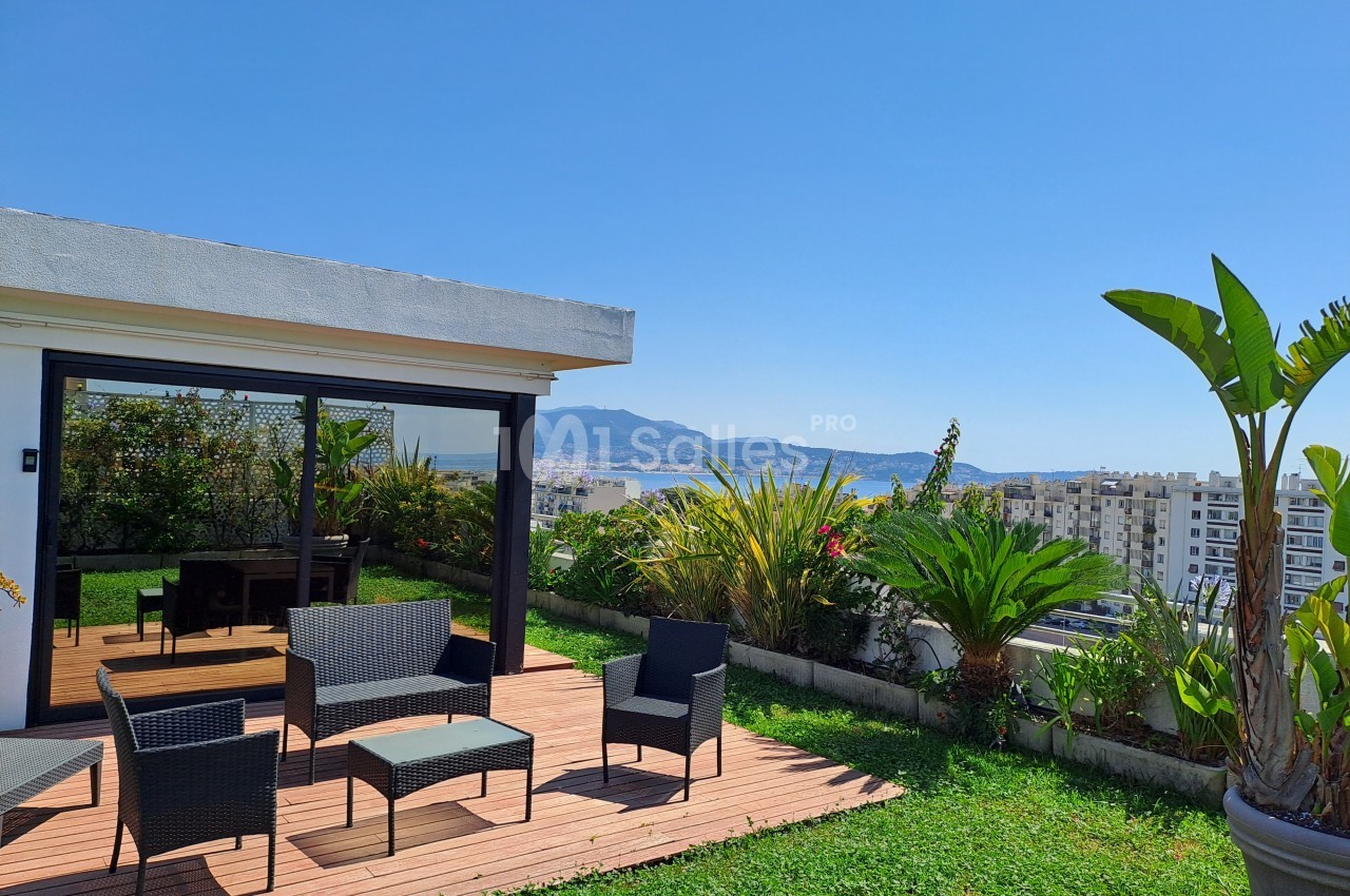 Terrasse avec mobilier en rotin, plantes, vue sur la ville, la mer et les montagnes sous un ciel dégagé.