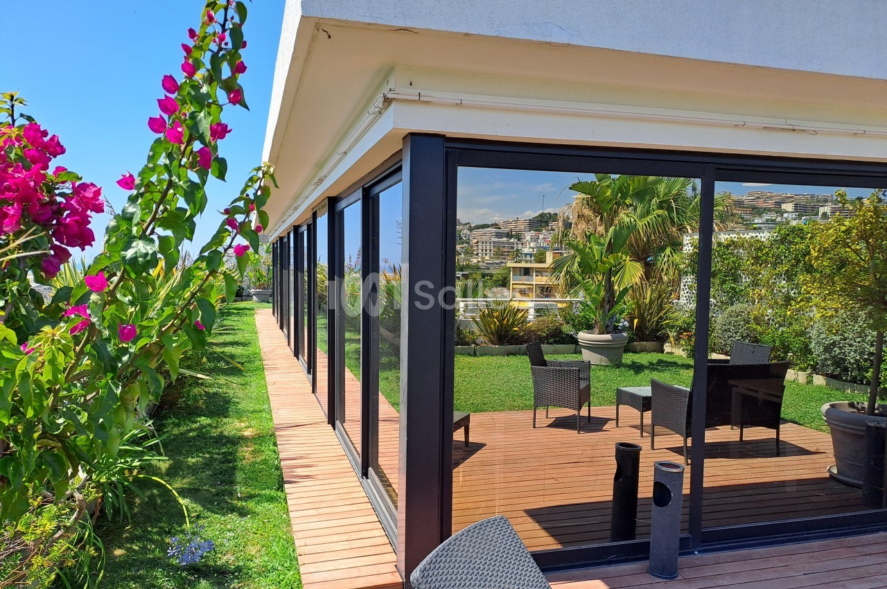 Terrasse en bois avec mobilier extérieur, entourée de verdure et de fleurs, jouxtant une maison aux grandes baies vitrées.