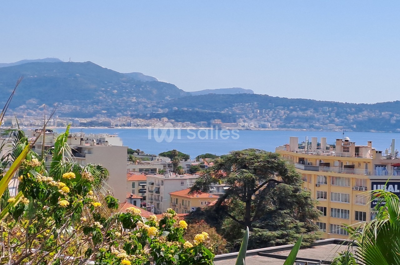 Vue sur une ville côtière avec des bâtiments, des arbres et des montagnes en arrière-plan, bordée par la mer.