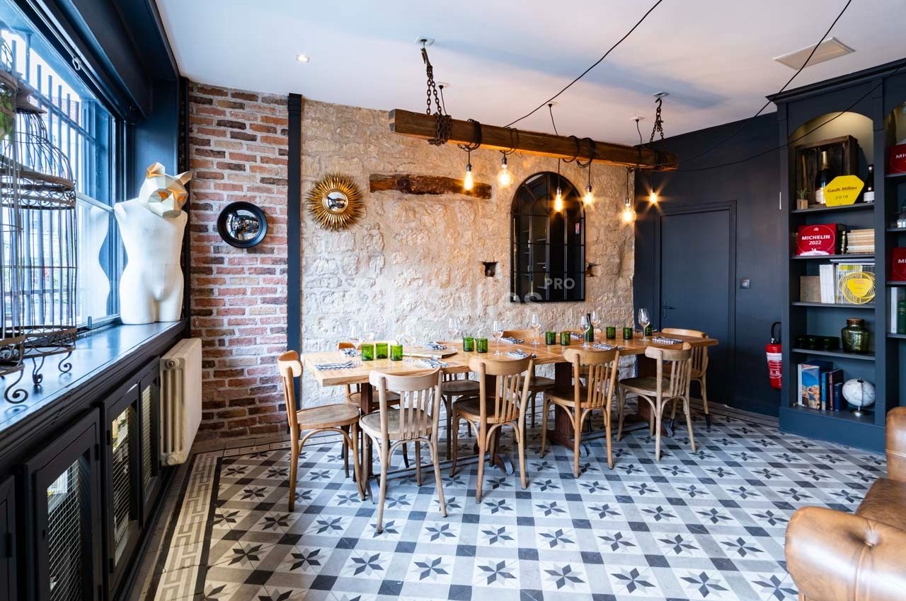 Salle à manger avec table en bois dressée, murs en pierre et briques, éclairage suspendu et sol à motifs géométriques.
