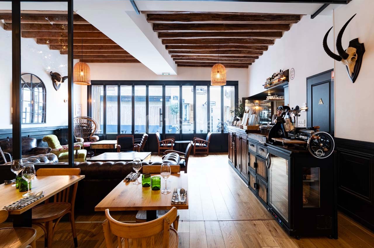Salle de restaurant lumineuse avec mobilier en bois, grandes baies vitrées et décoration murale éclectique.