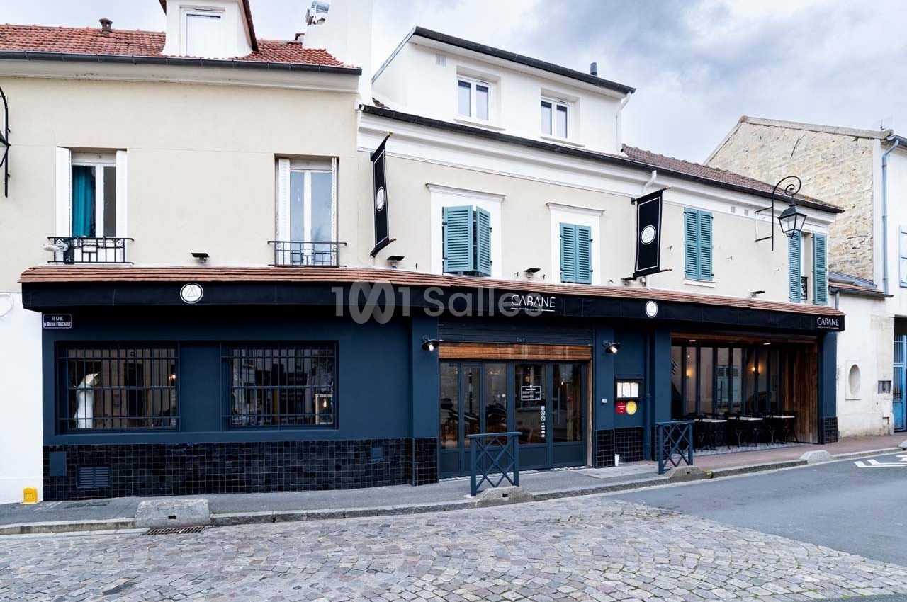 Façade d'un restaurant moderne avec murs bleu foncé, fenêtres vitrées et enseignes noires, situé dans une rue pavée.