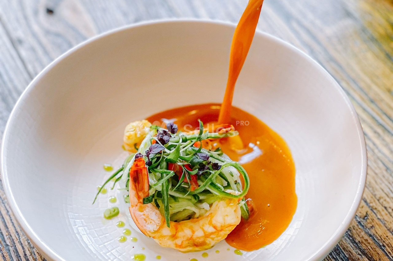 Crevette garnie de légumes verts et herbes fraîches, nappée de sauce orange dans une assiette blanche.