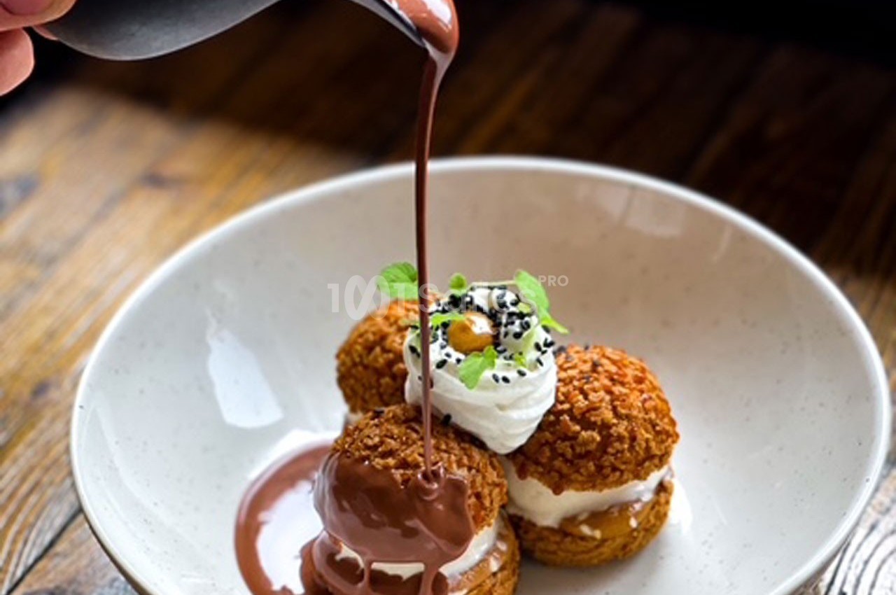 Des choux garnis de crème sont nappés de sauce chocolat, servis dans une assiette blanche sur une table en bois.