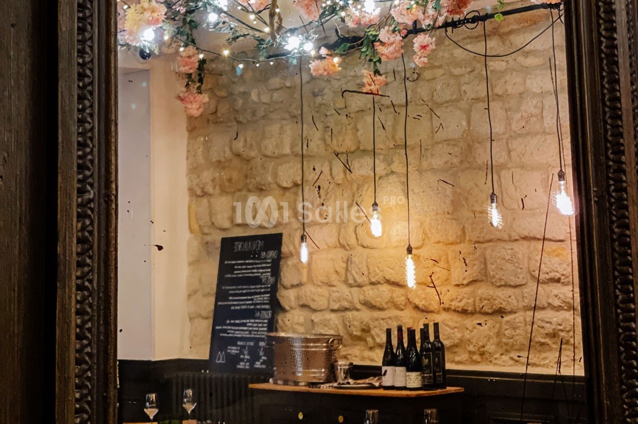 Salle de restaurant avec mur en pierre, guirlande lumineuse, bouteilles de vin et décor floral suspendu.