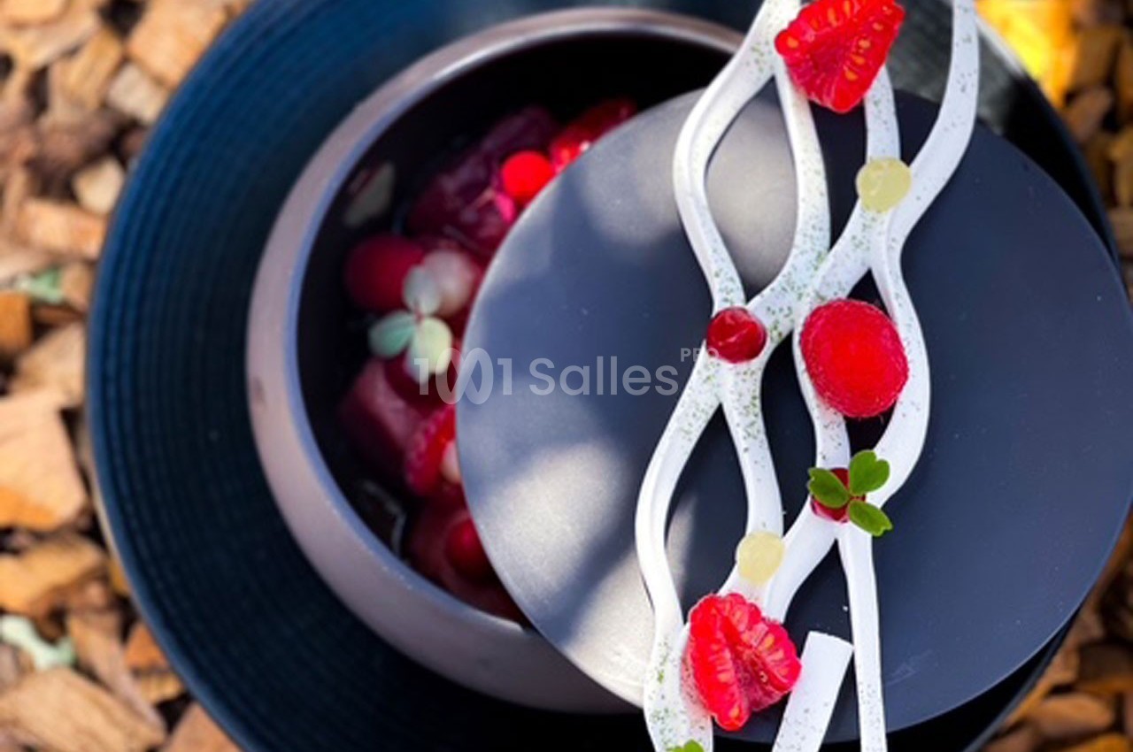 Dessert raffiné présenté dans un bol noir, décoré de fruits rouges, de crème blanche et de touches végétales.