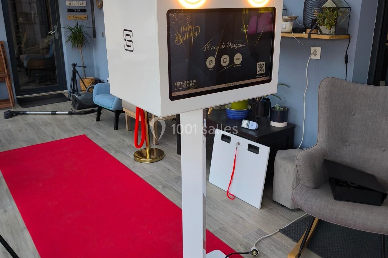 Borne photo interactive blanche sur pied, équipée de deux lumières circulaires, installée sur un tapis rouge dans un espace…