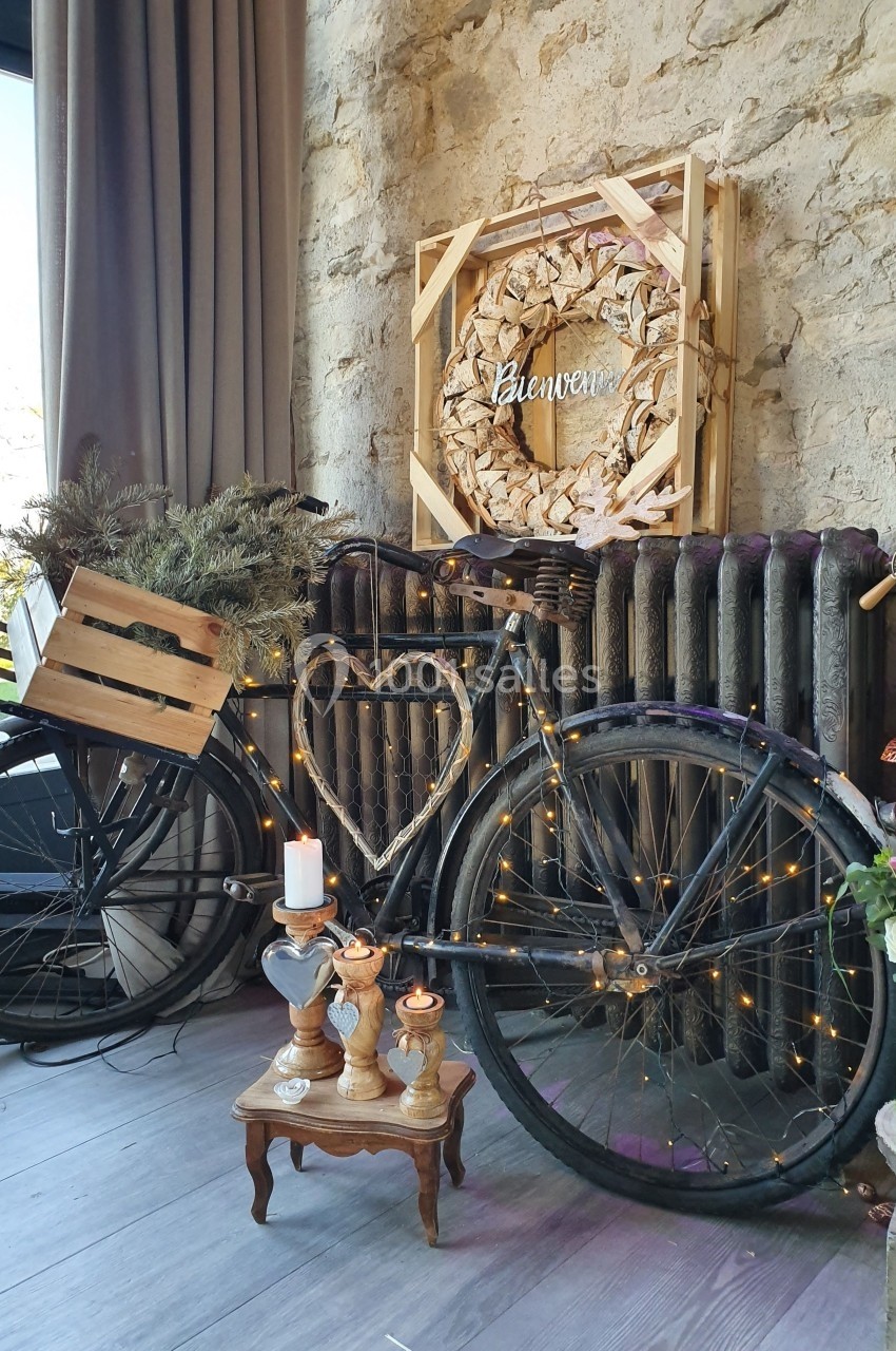 Vélo ancien décoré de guirlandes lumineuses, posé contre un mur en pierre avec une couronne et des bougies.