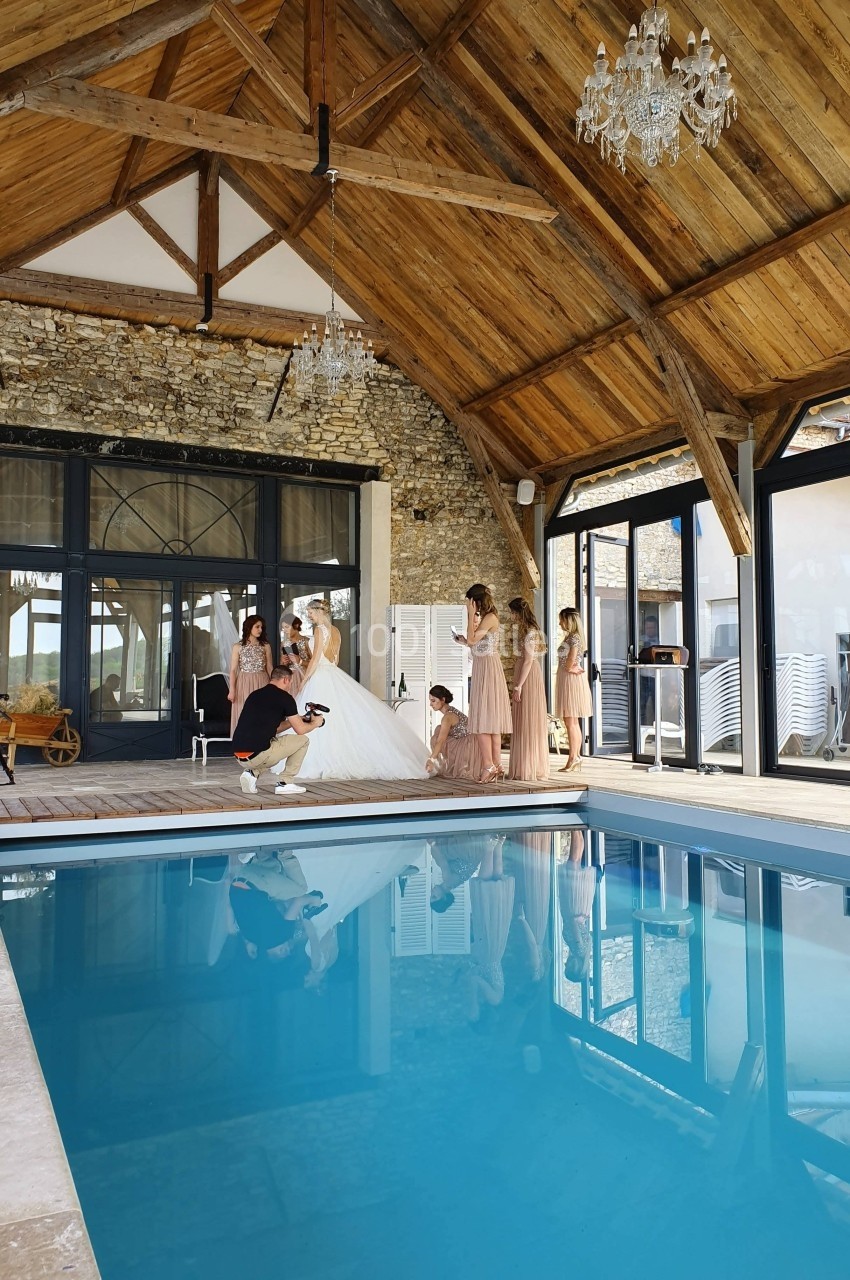 Mariée entourée de demoiselles d'honneur dans une salle avec piscine intérieure, poutres en bois et murs en pierre.