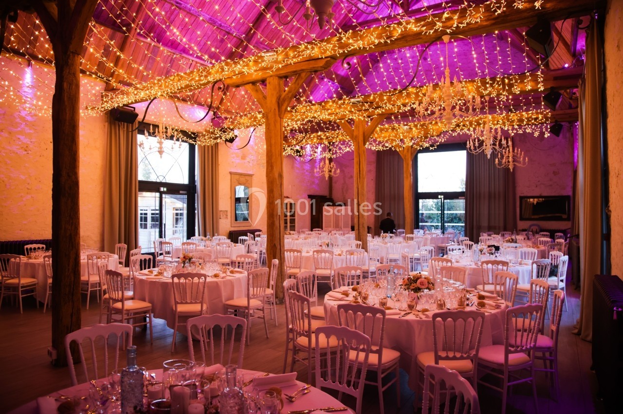 Salle de réception décorée avec des guirlandes lumineuses, des tables rondes dressées et des chaises blanches.