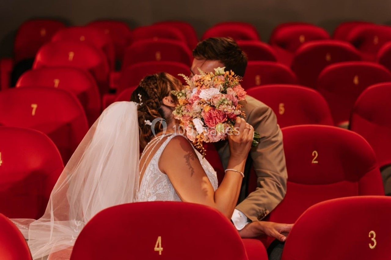 Un couple de mariés s'embrasse discrètement dans une salle de cinéma, entouré de fauteuils rouges numérotés.