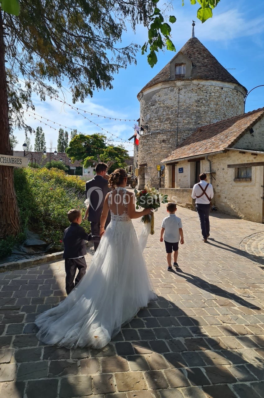 Une mariée en robe blanche marche sur une allée pavée, accompagnée de deux enfants et un homme, près d'un bâtiment en pierre.