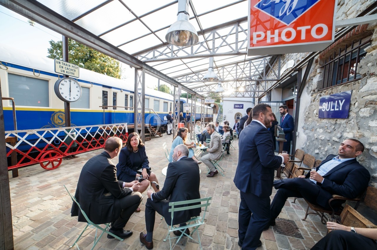 Personnes discutant en extérieur dans une gare ancienne avec des trains bleus et blancs en arrière-plan.
