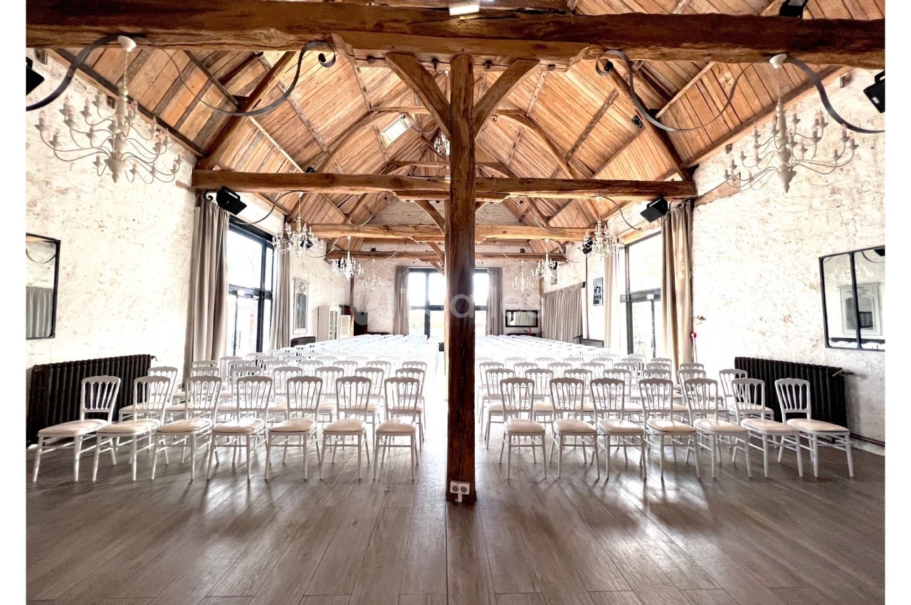 Salle lumineuse avec charpente en bois apparente, rangées de chaises blanches alignées pour un événement ou une réunion.