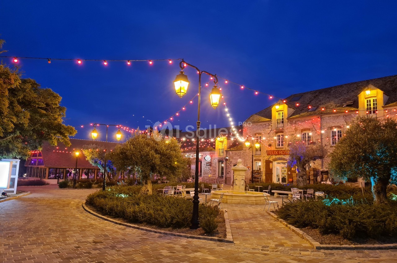 Place pavée illuminée par des guirlandes lumineuses et des lampadaires, entourée de bâtiments en pierre au crépuscule.