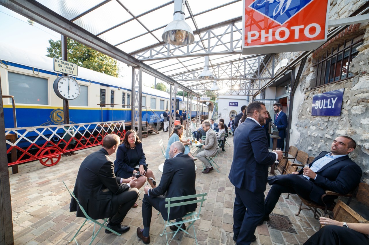 Personnes discutant en petits groupes sur une terrasse pavée près d'un train bleu et blanc dans une ambiance conviviale.