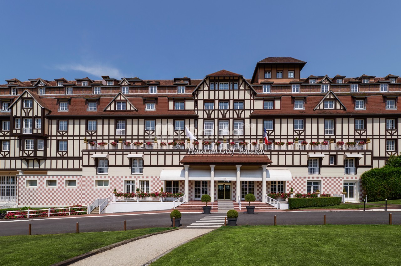 Façade d'un grand bâtiment à colombages avec balcons fleuris, entouré d'une pelouse et d'un chemin pavé.