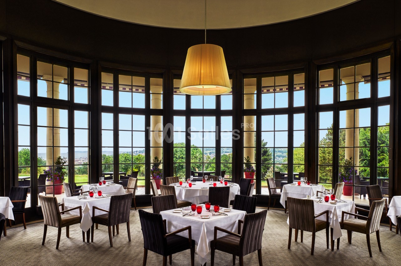 Salle de restaurant élégante avec grandes baies vitrées, tables dressées et vue sur un paysage verdoyant.
