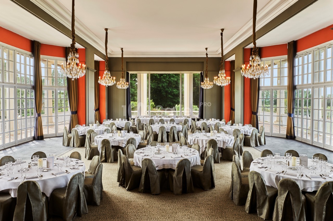 Salle de réception élégante avec tables rondes dressées, chaises couvertes et lustres, donnant sur un jardin verdoyant.