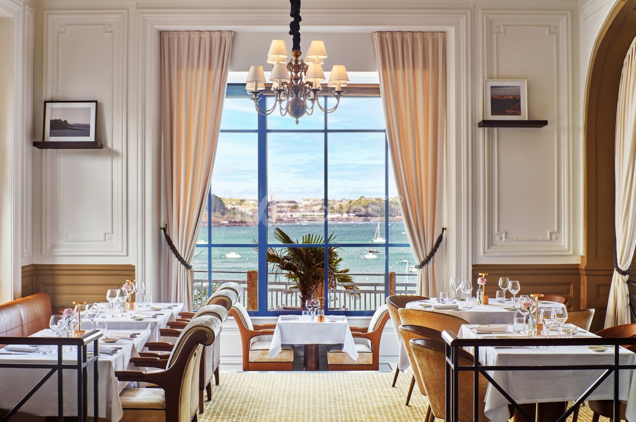 Salle de restaurant élégante avec vue sur la mer, grandes fenêtres, tables dressées et décoration raffinée.